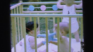 1964: three babies in a crib outdoors and two are playing with a toy