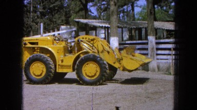 1965: a view of huge yellow tractor sitting in front of a barn and a white fence