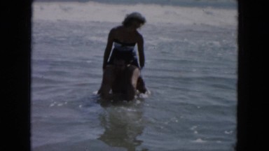 1961: man wading in waist high water at the beach with woman on his shoulders