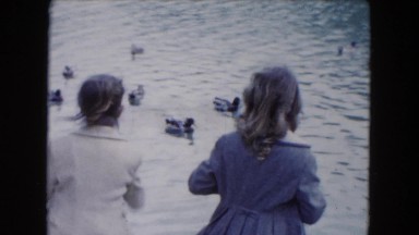 1961: ducks paddle in the water and eat food thrown by two girls standing by
