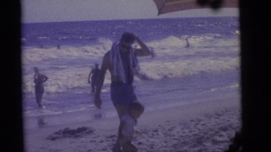 1964: man encourages child to wade on the beach, others play in the background
