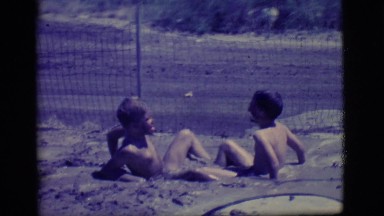1968: two young boys playing in a muddy, water filled hole in the ground.