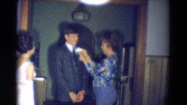1969: a mature woman pins a corsage to the suit of a young male