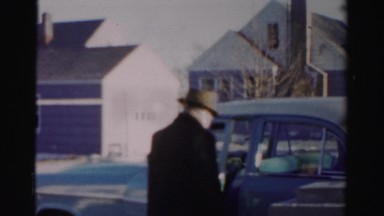 1956: a man gets into his car and moves the car toward a large garage and parks