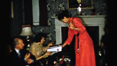 1956: a lady wearing a red robe is offering chocolates to her guests