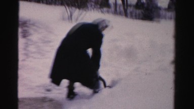 1956: a woman wearing a black coat and a scarf is vigorously shoveling snow