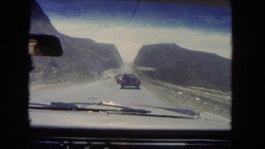 1962: windshield view of many cars driving on road through a mountain range