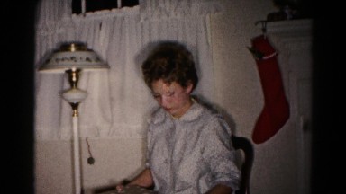 1959: woman in nightgown opening christmas present next to lamp and stocking