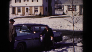 1959: elderly lady carrying packages walks gingerly through the snow past a car