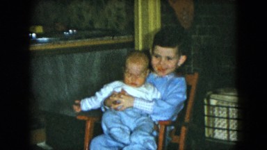 1951: a boy wearing blue is holding his baby brother in a chair in the living