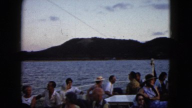 1962: passengers relax along a railing on ship moving near shoreline