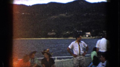 1962: men and women sail on the water with houses and mountains in the view.