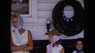 1961: a woman and two girls wearing hats with scarves outdoors along a wall