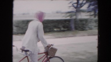 1961: a woman with scarf riding a bike behind several horse-drawn carriages
