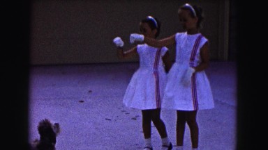 1961: two girls dressed in the same dress play with a small dog in the driveway