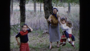 1955: kids playing outdoor with their mother next to a blue shake shingle house