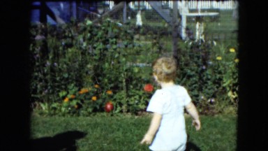 1951: an adult off-camera is blowing bubbles for a toddler boy in the backyard