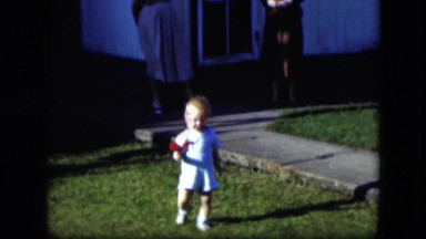 1951: a cute little toddler walking through the yard holding a red flower