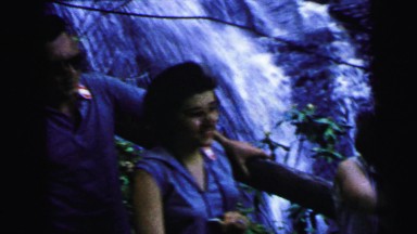 1964: two women and a man standing by a waterfall waving their hands. CAMDEN