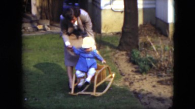 1951: baby try to stay on a little rocking chair with the help of an adult woman