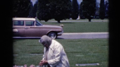 1968: a woman kneeling alone at a grave site with a car in the background