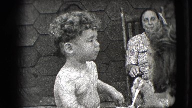 1942: shirtless little toddler is being spoon fed by his mother