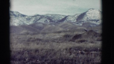 1964: grassy field sits at the base of snow covered mountains on a cloudy day