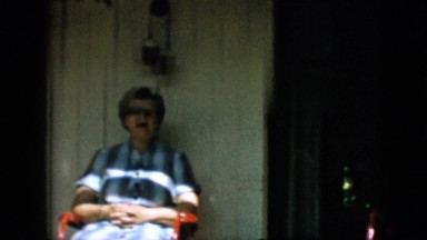 1957: older woman with a plaid dress sitting on a porch waving at the camera