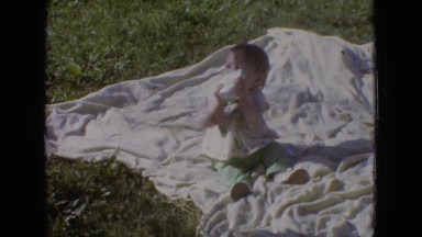 1965: a baby sits on a blanket in the grass, drinking from a paper cup