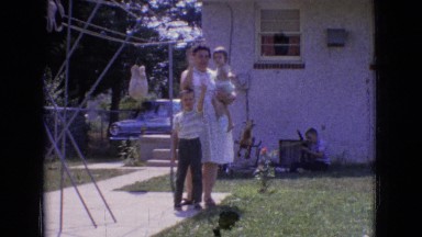 1965: a woman outside with a boy waving as she holds a baby in one arm