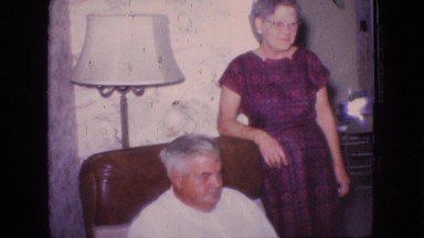1966: old man sitting on a sofa like chair and wife along side standing