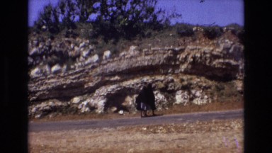1972: Two Ladies in Black Robes Walking Down Road in Hilly Area with Green Trees