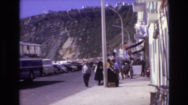 1972: pedestrians walking up street below a looming cliff against the sky