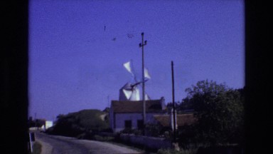 1972: white sails of a windmill turn behind a house and small road against blue