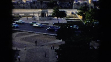 1956: cars drive on busy city street bordering sidewalk and fountain with statue