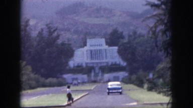 1961: car moves towards white large bungalow midst of greenery and mountain