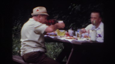 1967: two men and a lady sharing a meal at a picnic table outside near a tree