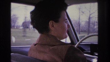1966: a white woman with dark brown hair in a brown suede jacket operates a car