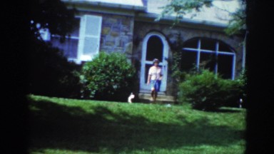 1962: lady coming out house walking towards garden trees round greenery puppy