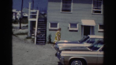 1962: older lady in yellow print dress walks past cars towards small house
