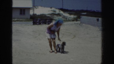1962: woman training her poodle to obey and sit while they are at the beach