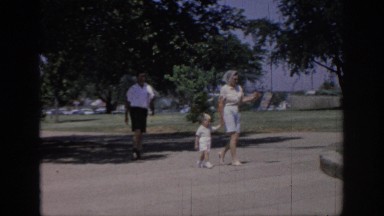 1967: mother holding child walking park father coming behind cars parked