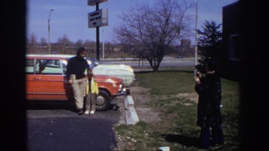 1973: woman taking a picture of man and child in front of an orange car