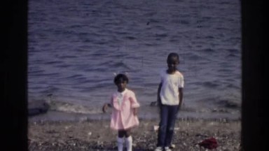 1976: school aged black brother and sister playing at a pebbled beach