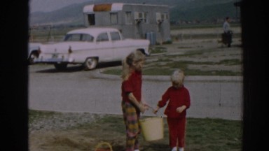 1962: two children in red shirts carry a bucket together and smile at the camera