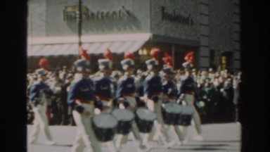 1962: marching band parades down the street whilst people watch on the side