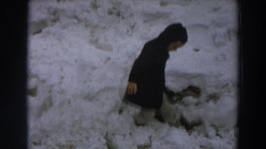 1958: child playing in the snow along a gravel road while parent rides a horse.