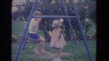 1965: two children playing on blue swingset and pushing each other on summer day