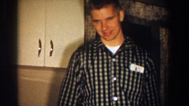 1958: Man in Plaid Shirt and Glasses Walking with Arms Crossed While Conversing with Woman in Home Setting