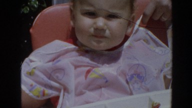 1964: child sitting in highchair with a bib being fed food with a spoon
